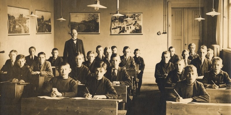 Ett äldre klassrum med elever i bänkarna sett framifrån. Läraren bakom. Skolplanscher på väggarna.