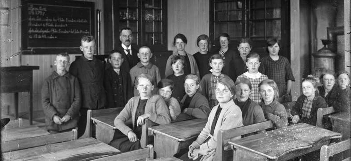 Äldre gruppfoto av en grupp elever och en lärare i skolsal med värmekamin i bakgrunden.