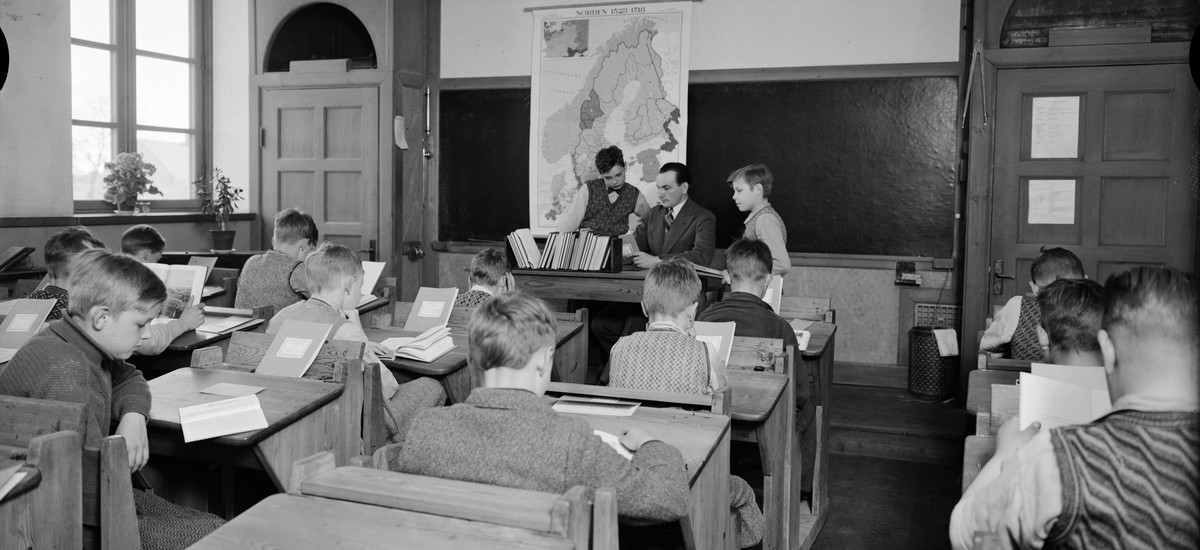 Ett klassrum med elever vid sina bänkar, undervisning pågår, magister samtalar med två elever vid katedern