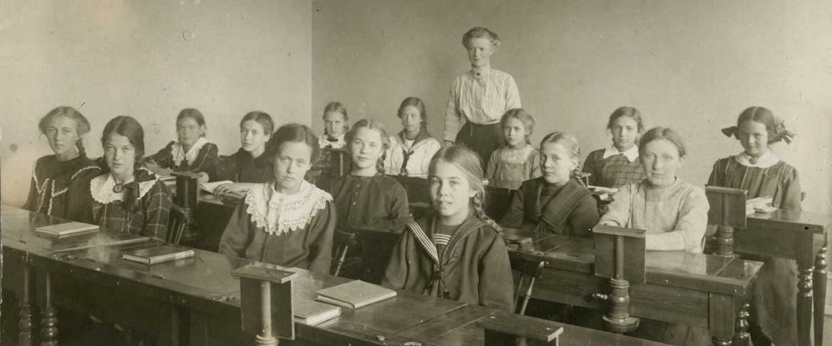 Flickor vid sina bänkar med lärarinnan längst ner i klassrummet.